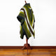 A striped, multicolored poncho is draped on a wooden mannequin stand; it features green, white, and brown hues, positioned in a room with wooden flooring and a plain white wall.