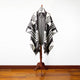 A poncho with black and white geometric patterns is displayed on a wooden mannequin stand, set against a white wall and wooden floor.