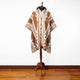 Poncho displayed on a wooden stand, featuring geometric patterns in brown and beige. It's set against a plain white wall, resting on a wooden floor.