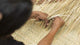 Hands weave dried leaves into a basket on a textured mat, demonstrating traditional crafting techniques in a natural setting.
