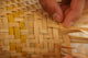 Hands weave light and dark straw strips into a textured pattern, creating a basket. The close-up focuses on the intricate interlacing technique, highlighting craftsmanship and precision.
